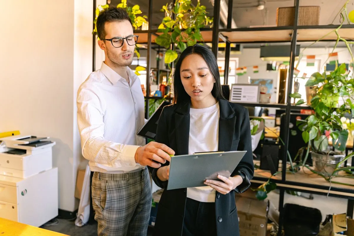 A man in glasses and a white shirt points at a clipboard held by a young woman in a black blazer as they stand talking in a bright office lined with shelves of hanging plants.
