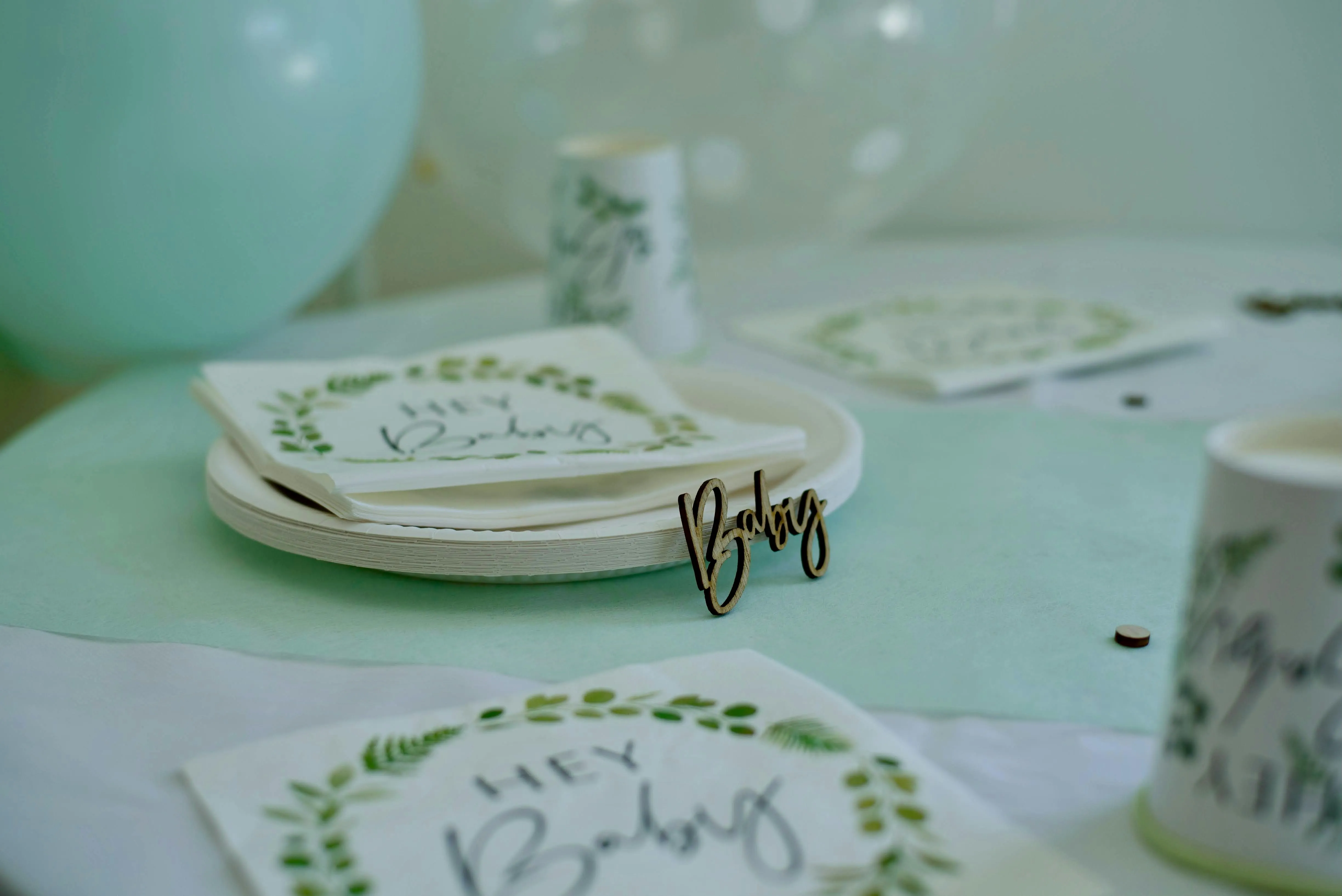 Pastel green baby shower table setting with 'Hey Baby' napkins, balloons, and a wooden 'Baby' sign on a mint tablecloth