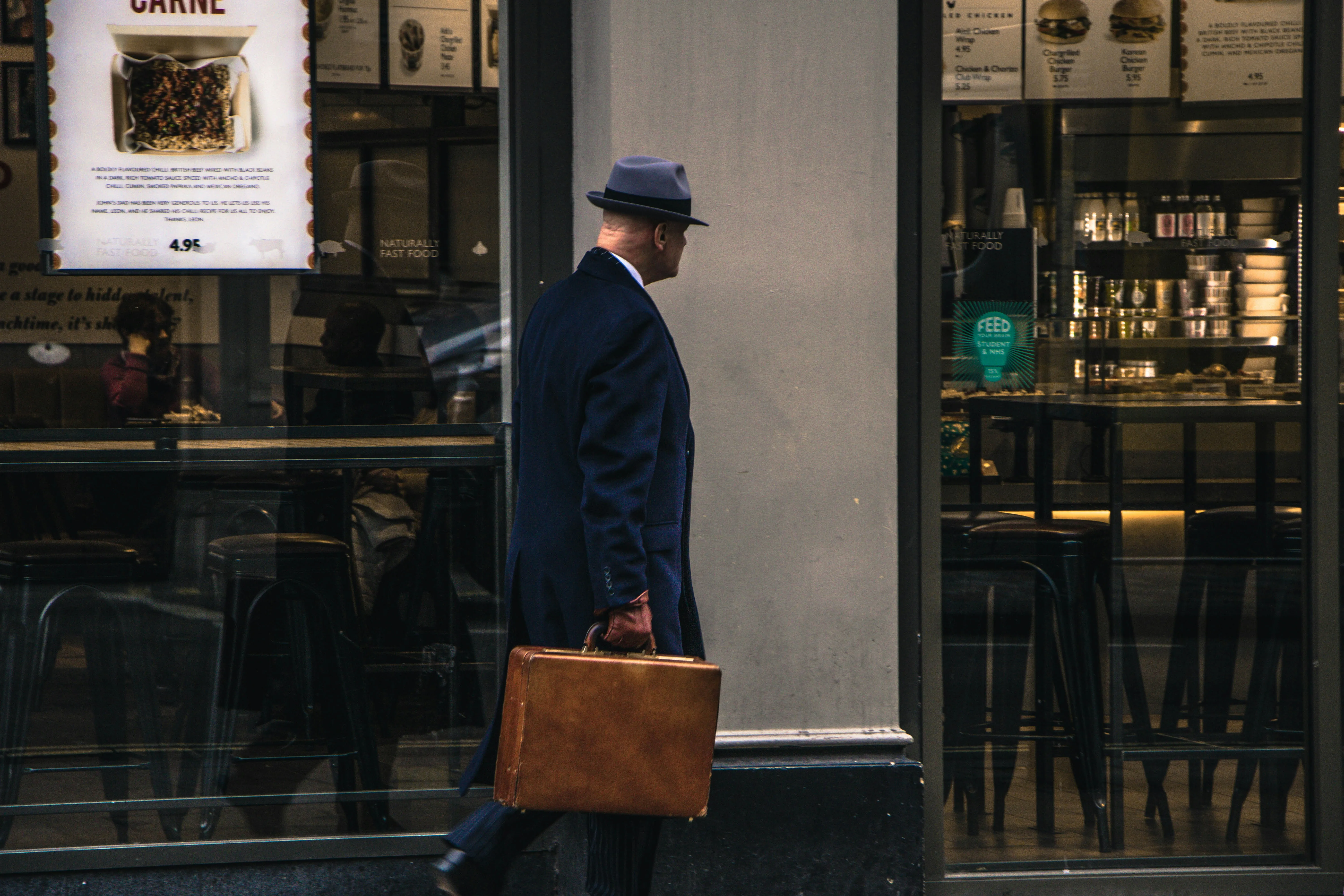 Man in a long dark trench coat and fedora carrying a leather briefcase past a city storefront, evoking classic spy-thriller imagery