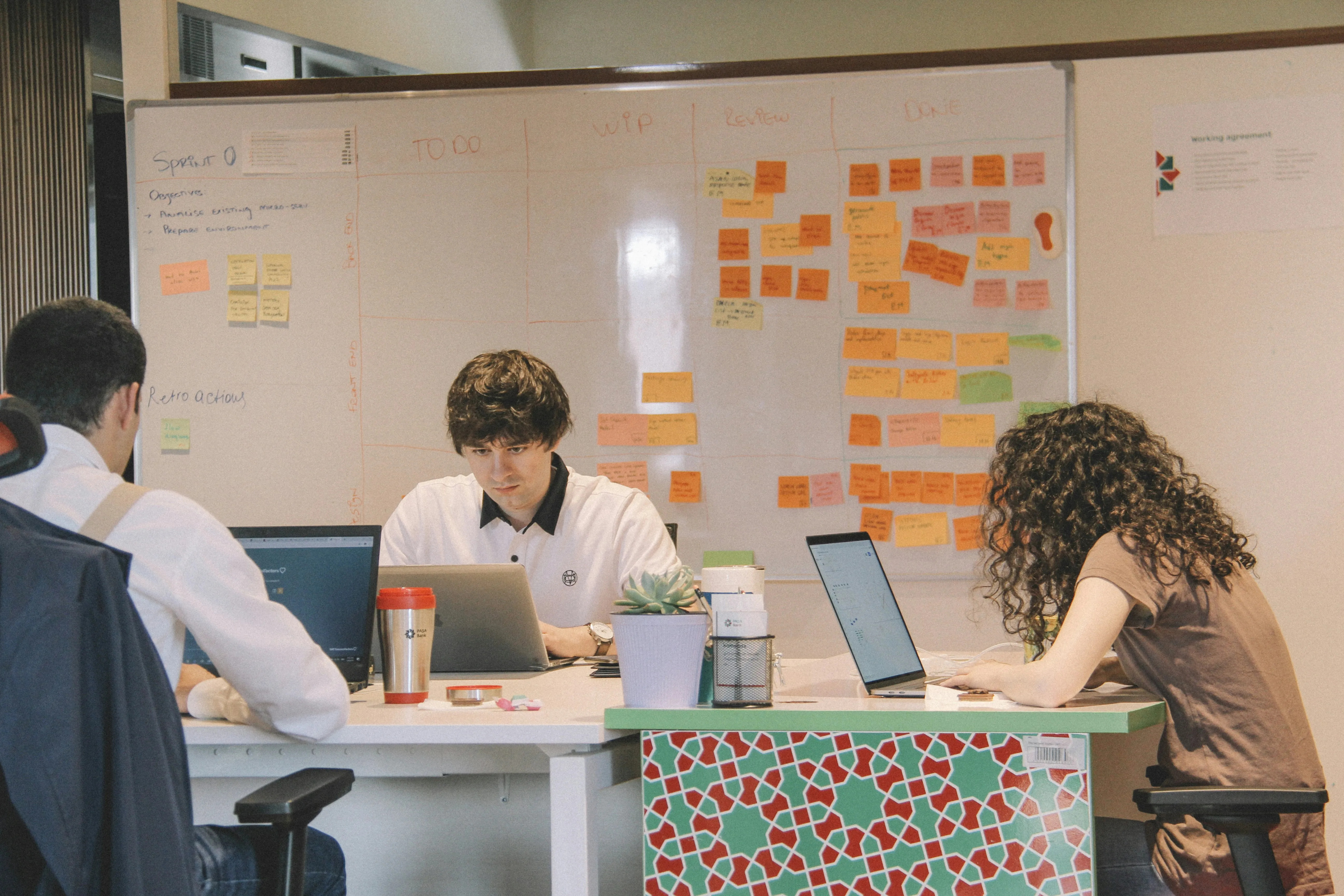 Three teammates working at desks in front of a whiteboard covered in sticky notes arranged into agile sprint columns