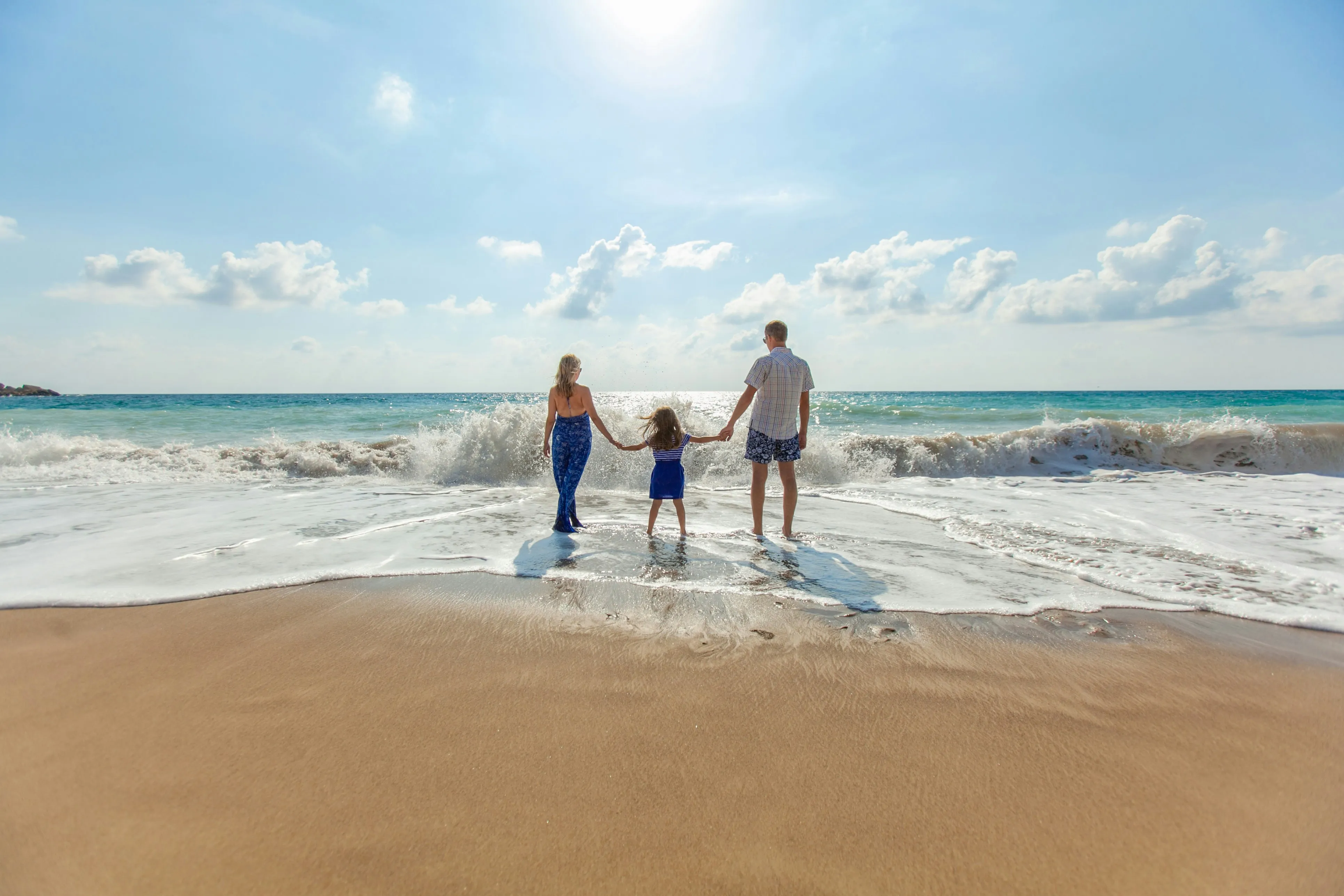 Family of three holding hands on a sunlit beach with gentle waves rolling in over the wet sand