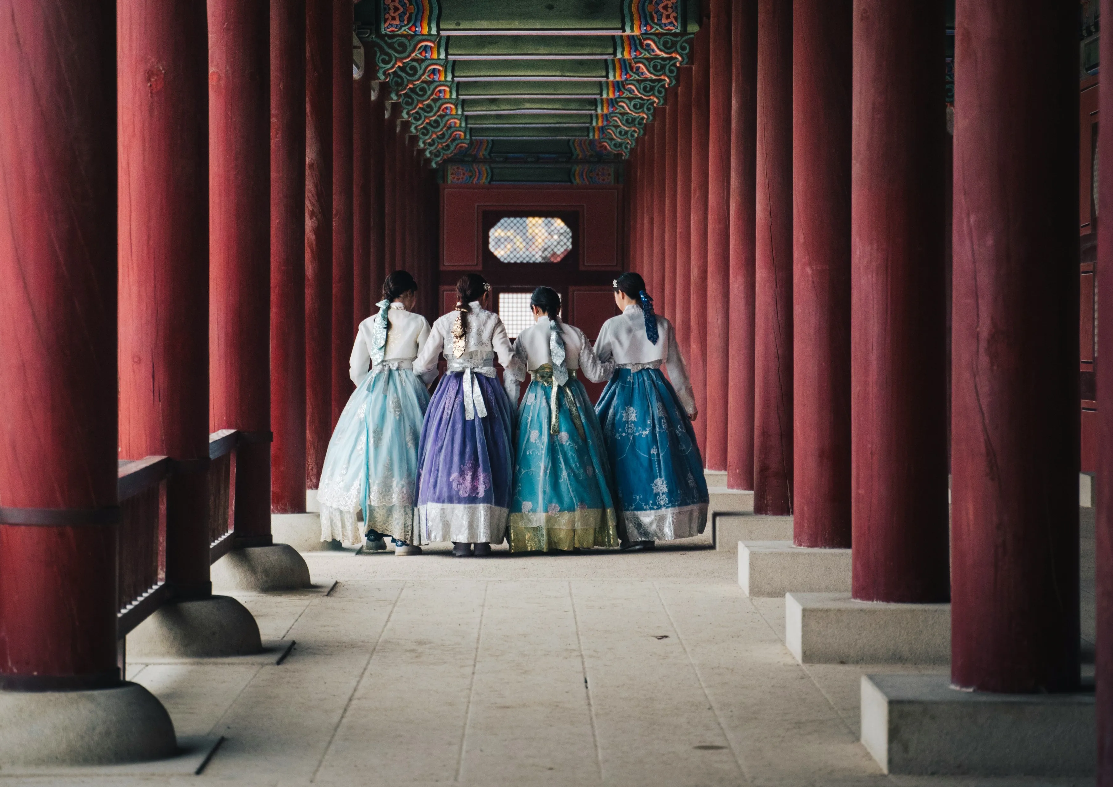 Four women in colorful traditional Korean hanbok dresses walking through a red-pillared corridor at a Korean palace