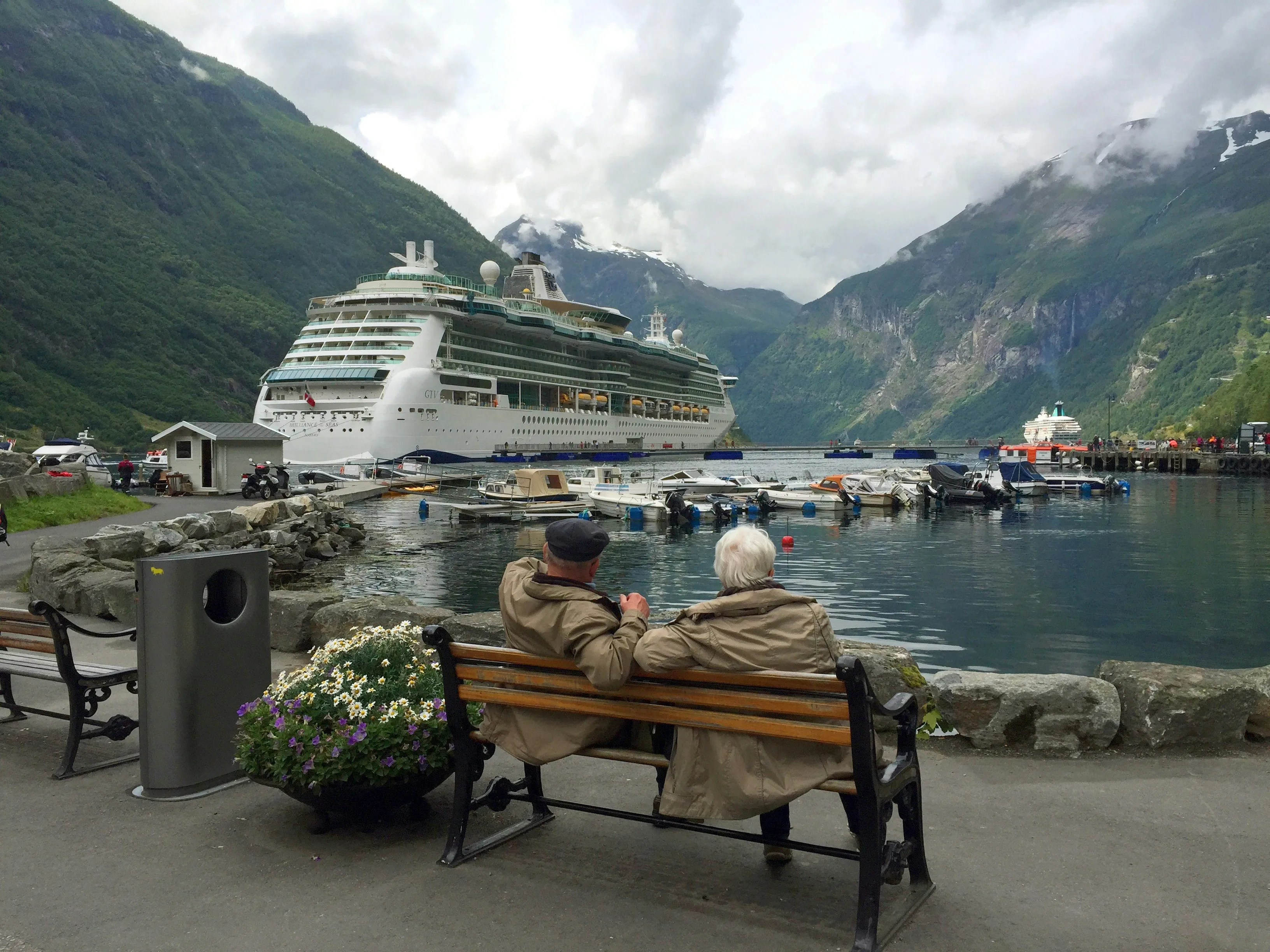 Older couple sitting on a wooden bench at a harbor, looking out at a docked cruise ship surrounded by green Norwegian fjord mountains