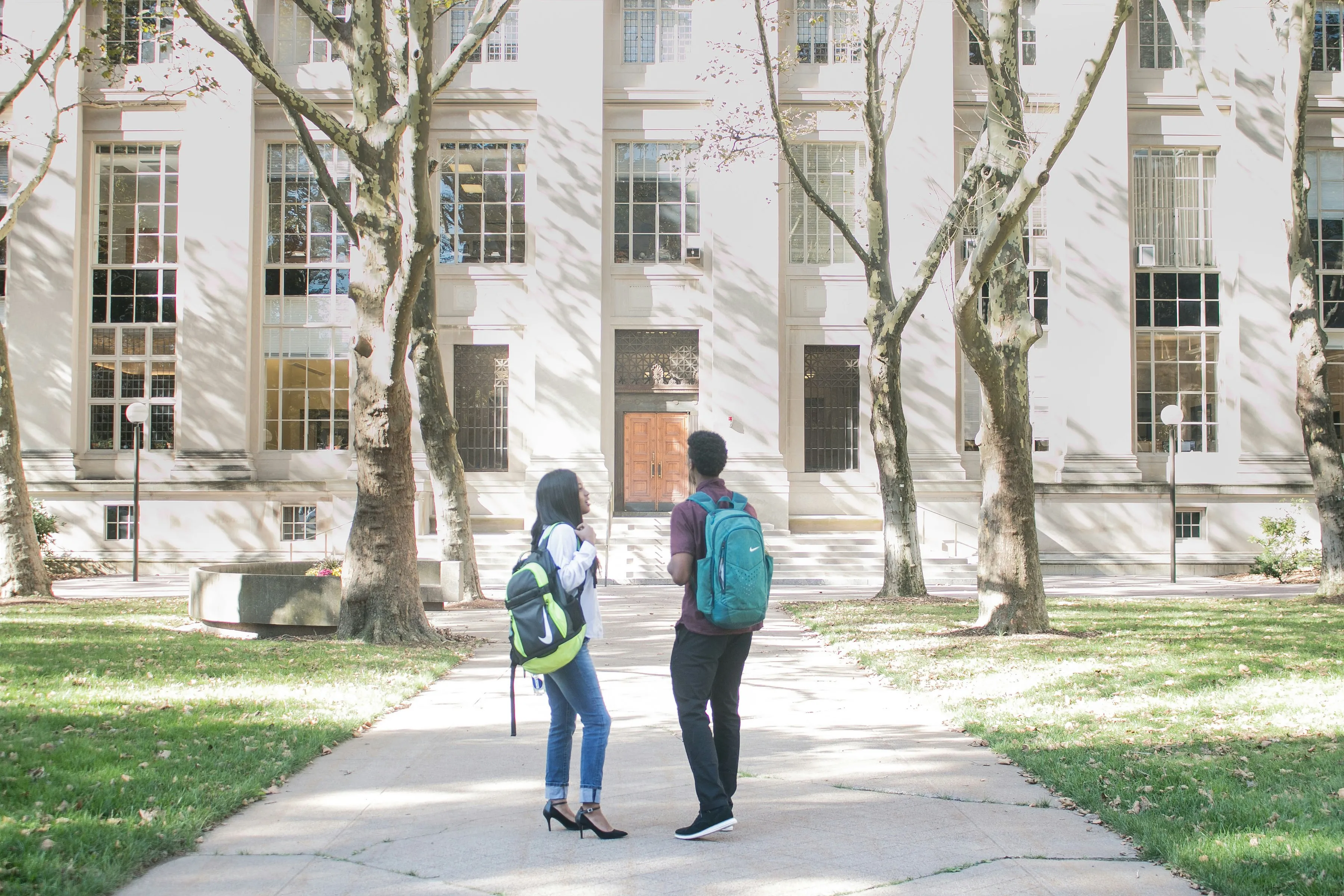 Two college students with backpacks standing on a sunlit campus walkway in front of a tall white academic building