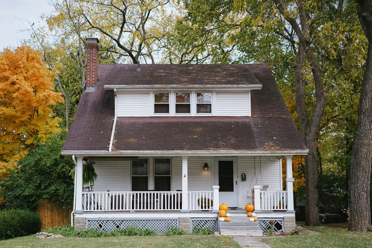 White suburban two-story house with a covered front porch and pumpkins on the steps, framed by autumn trees