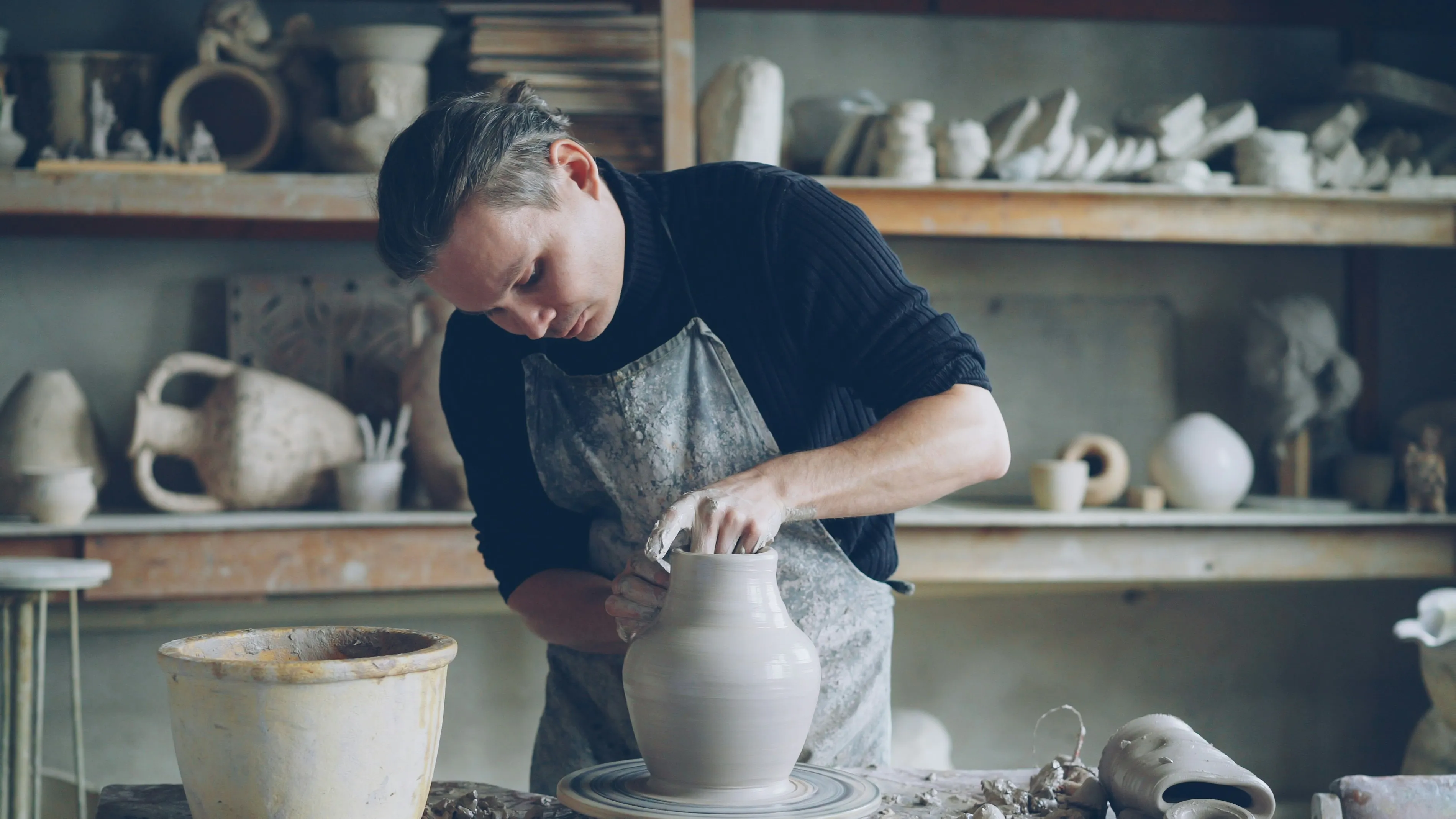 Potter in a clay-stained apron shaping a tall white vase on a pottery wheel inside a workshop full of finished ceramic pieces