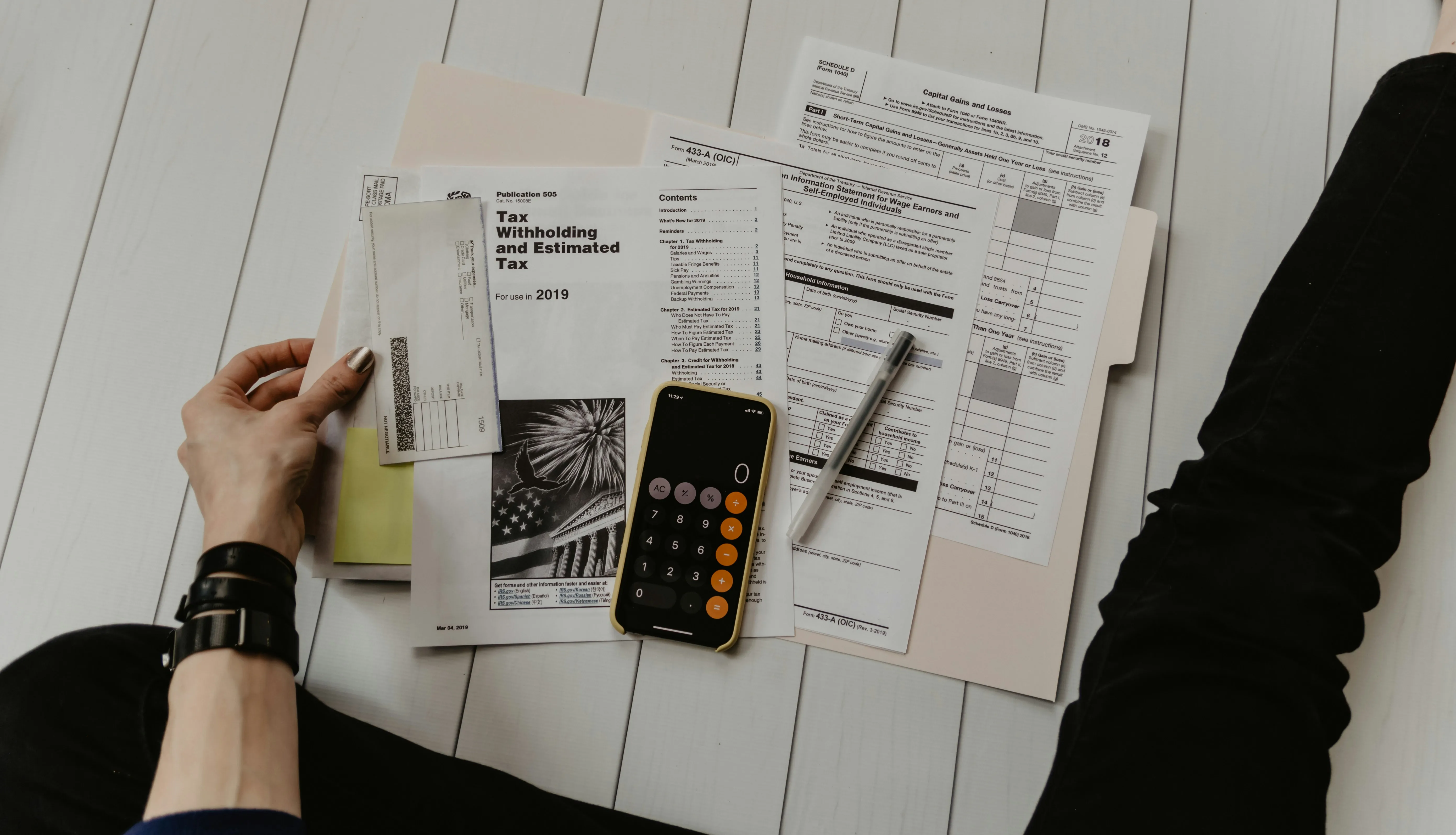 Overhead view of US tax forms, a calculator app open on a smartphone, and a pen spread across a white wooden desk
