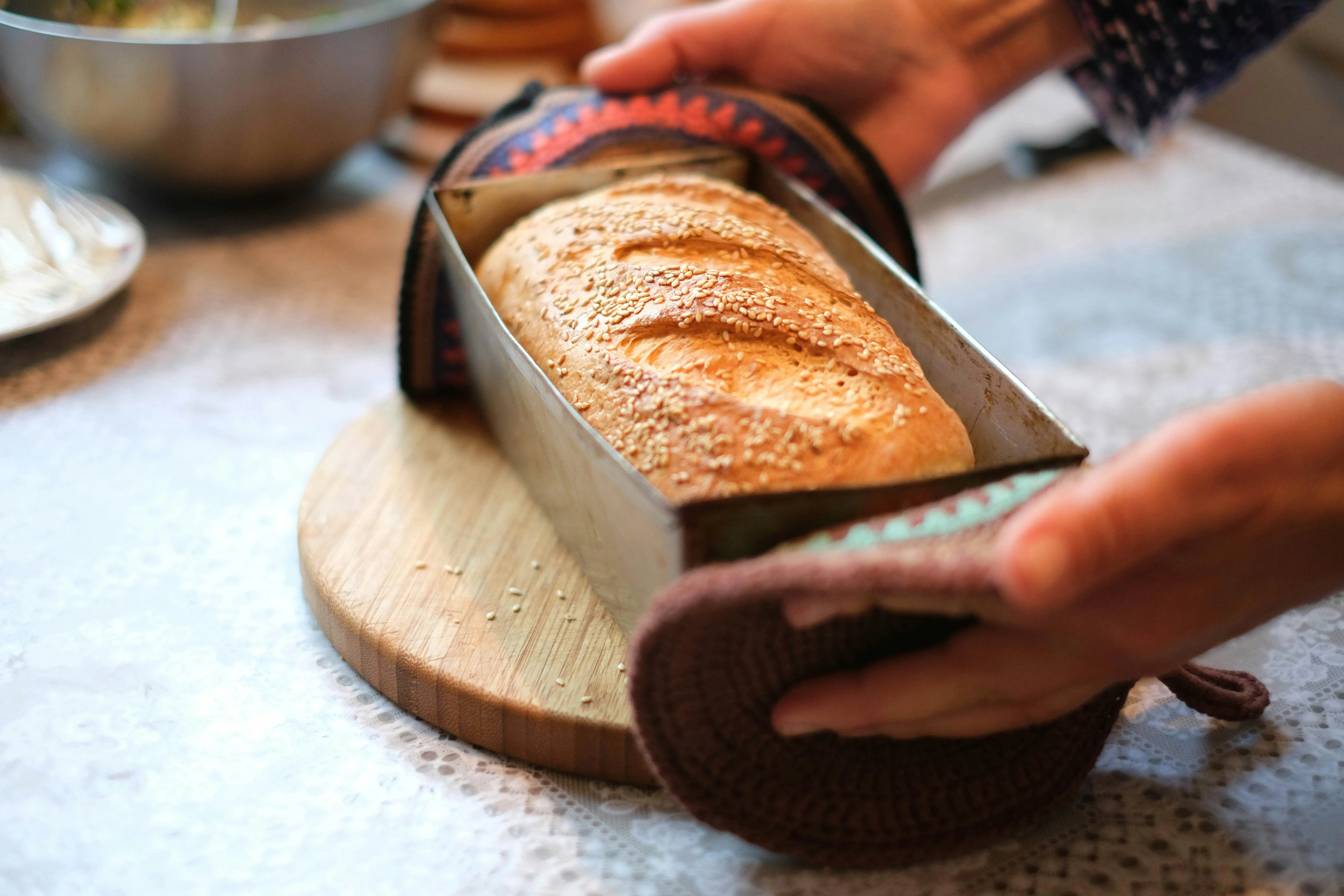 Hands in oven mitts lifting a freshly baked sesame-topped loaf out of a metal bread pan in a home kitchen