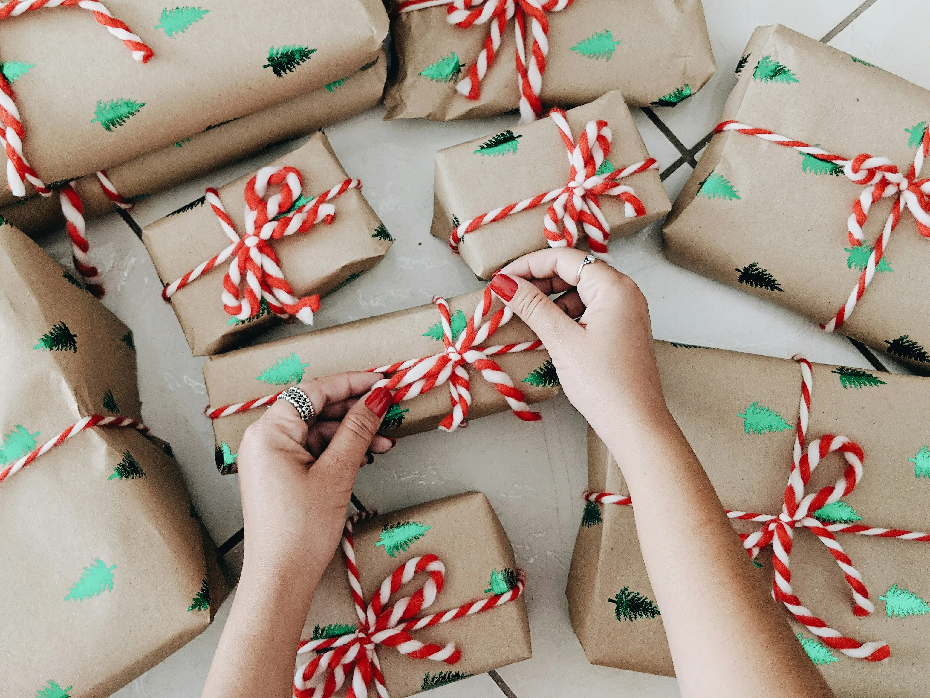 Hands tying a candy-cane-striped twine bow on holiday gifts wrapped in brown kraft paper printed with green Christmas trees