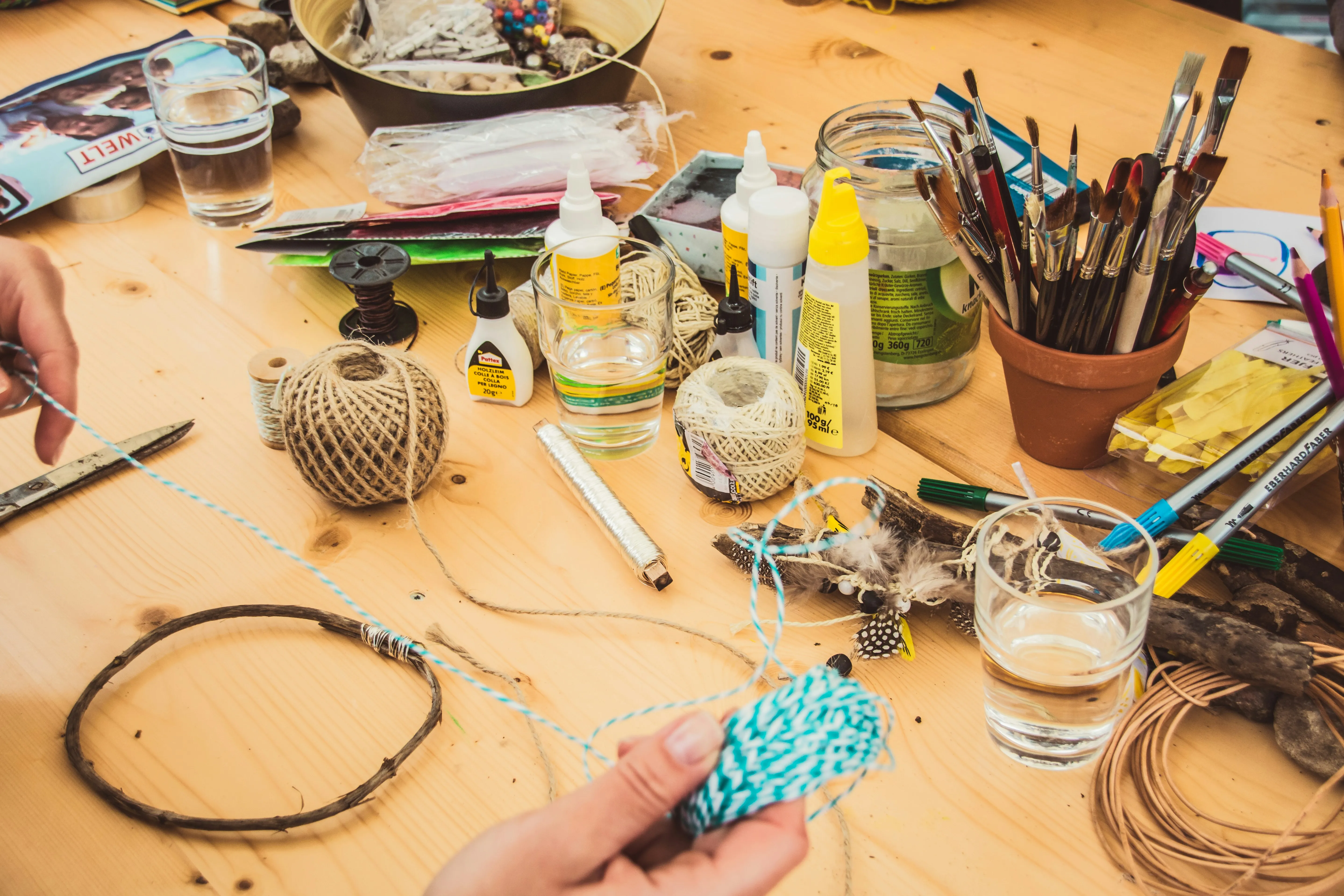 Wooden craft table covered in twine spools, glue bottles, paintbrushes, and beads with hands working on a beaded project