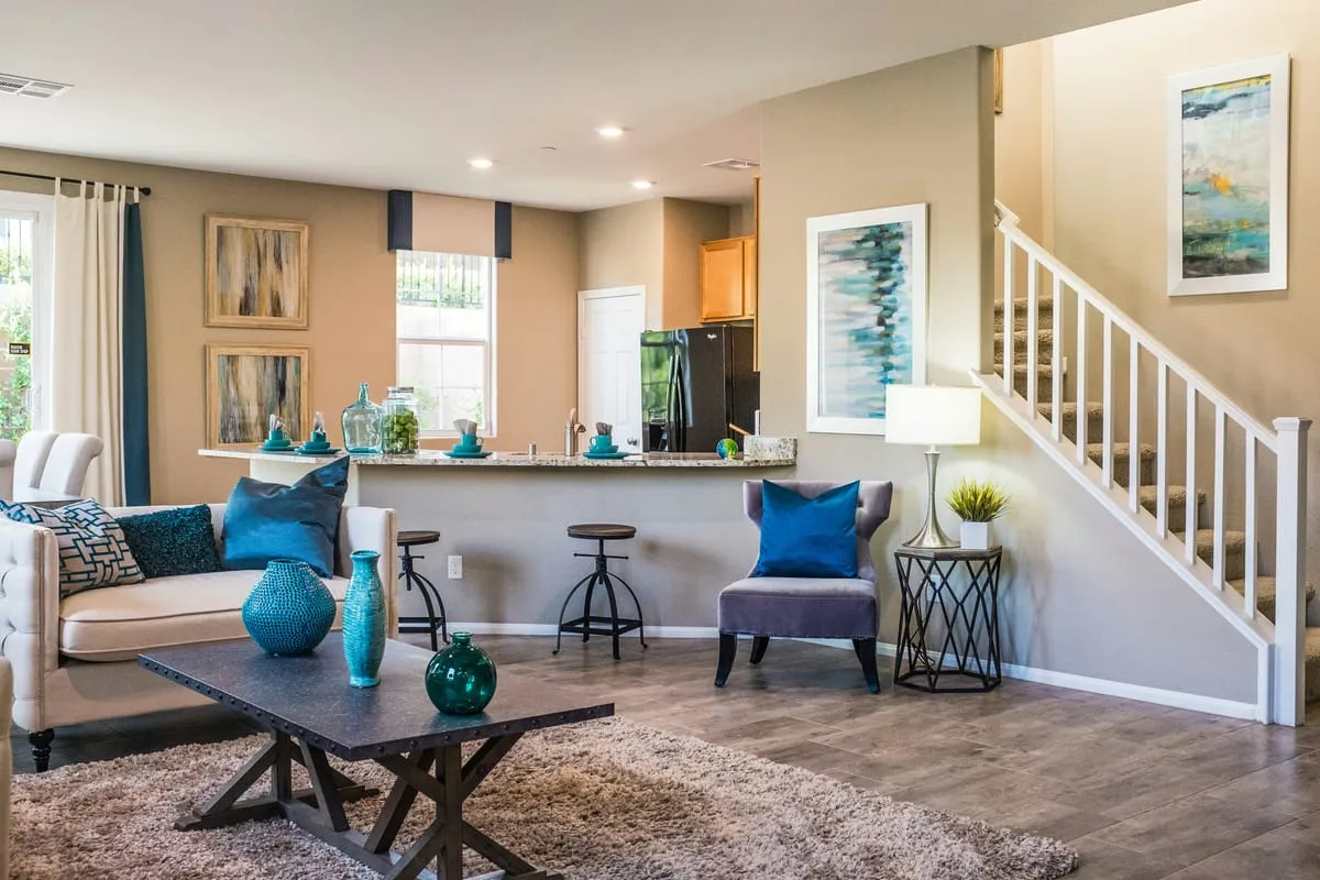 Bright open-concept living room with a beige sofa, teal accent decor, kitchen island, and a staircase visible in the background