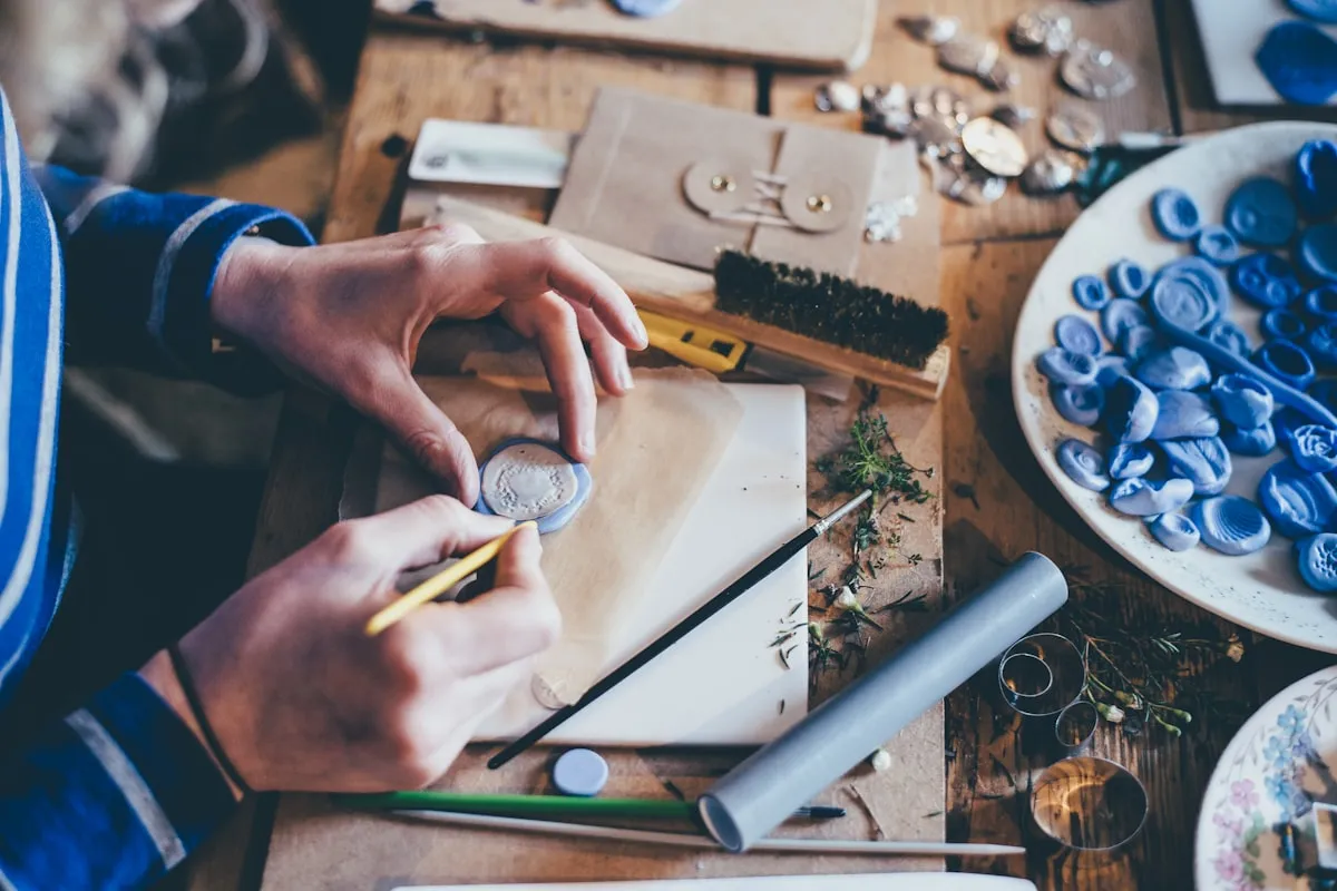 Overhead view of an artisan's hands shaping a wax seal at a wooden workbench scattered with tools, blue wax stamps, and crafting materials