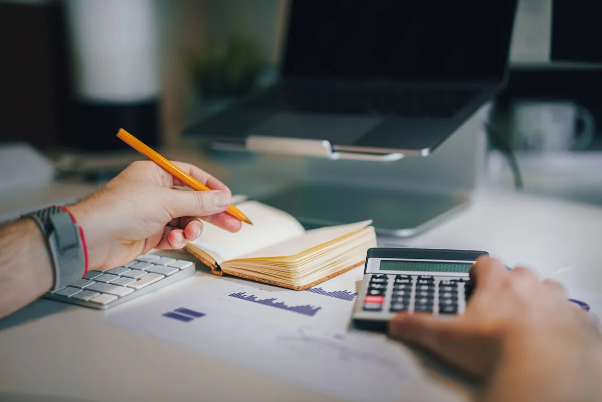 Hands at a desk reviewing finances — writing in a notebook, working a calculator, with a laptop and printed bar chart in view