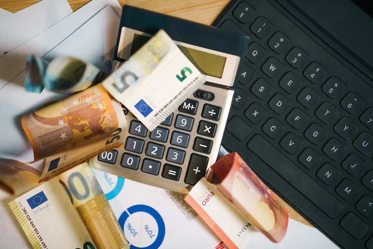 Calculator and laptop keyboard on a desk surrounded by scattered Euro bills, illustrating the costs and fees of running an online shop