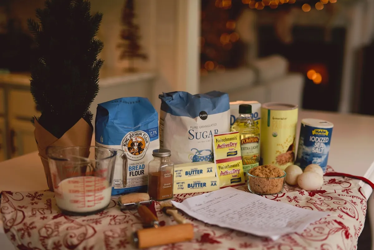 Baking ingredients arranged on a kitchen counter — bread flour, sugar, butter, yeast, eggs, and a handwritten recipe — with festive lights in the background
