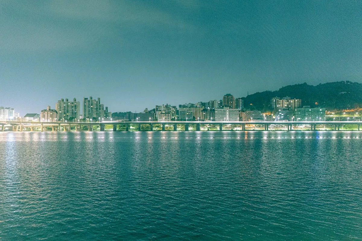 Seoul skyline at night reflected in the calm Han River, with city lights along the bridge spanning the water