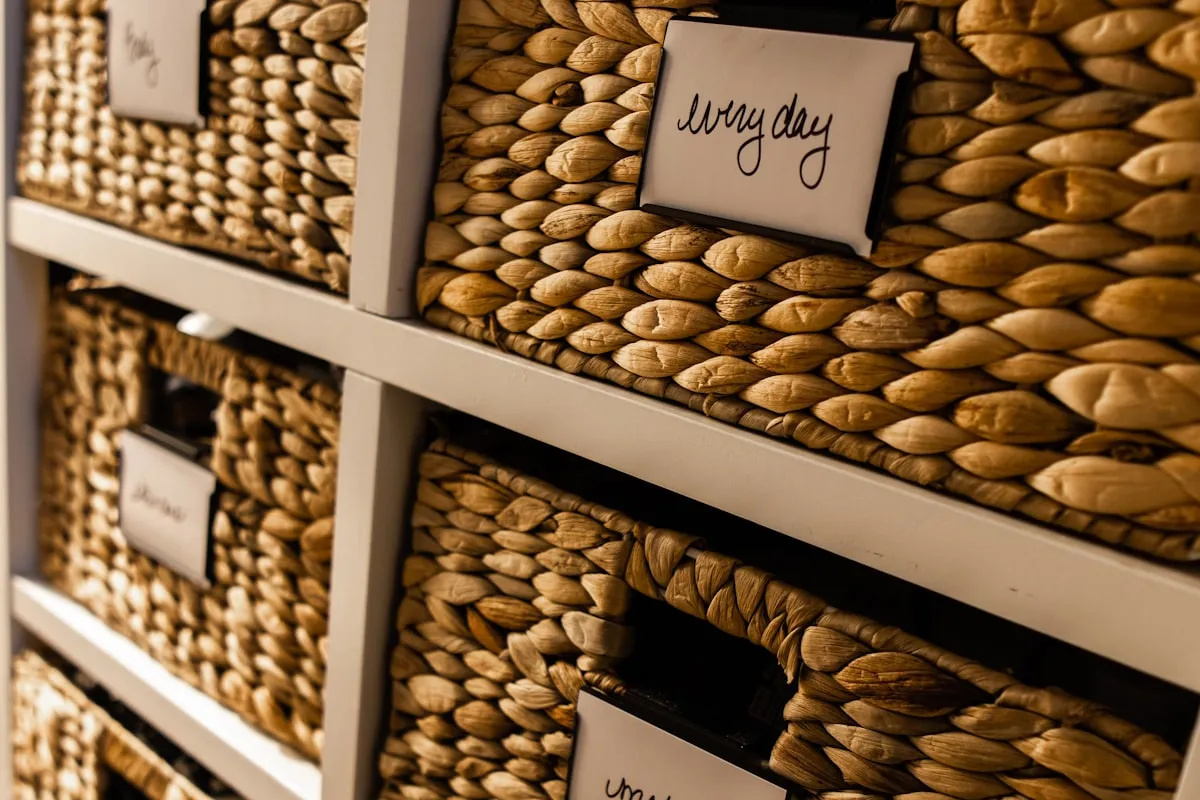 Close-up of woven wicker storage baskets in a shelving unit, each with a handwritten label card identifying its contents