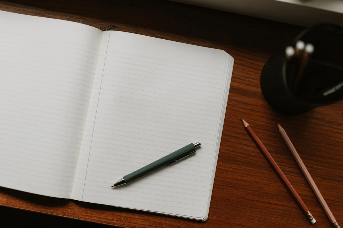 Open blank lined notebook and a green pen on a dark wood desk beside two pencils and a mesh pencil cup