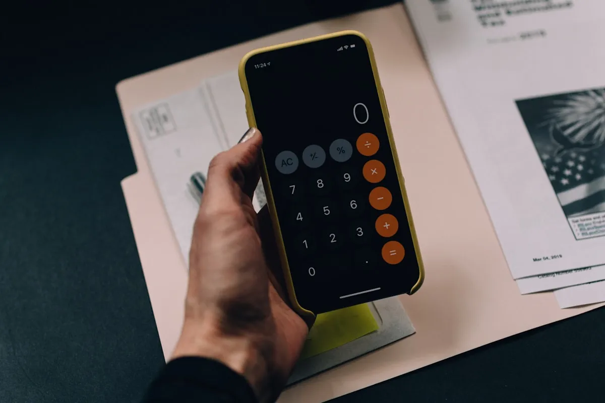 Hand holding a smartphone with the calculator app open above a manila folder of US tax documents on a dark desk