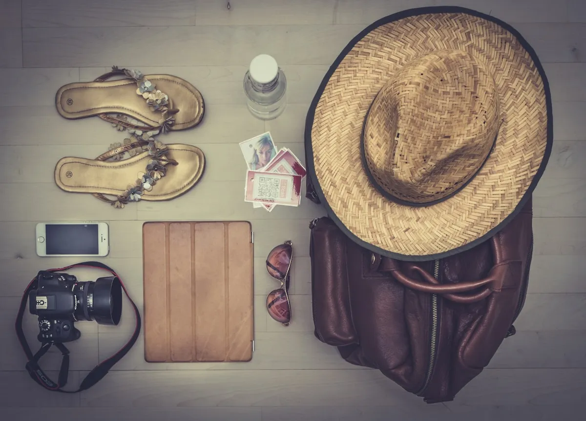 Flat-lay of travel essentials on a wood floor — straw sun hat, leather backpack, sandals, sunglasses, camera, smartphone, tablet, water bottle, and tickets