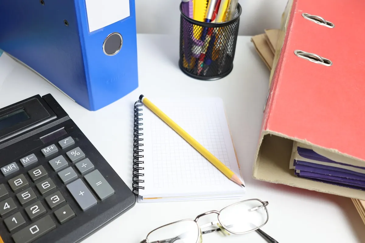 Calculator, blue and red ring binders, a notebook and pencil, a cup of supplies, and reading glasses arranged on a white desk