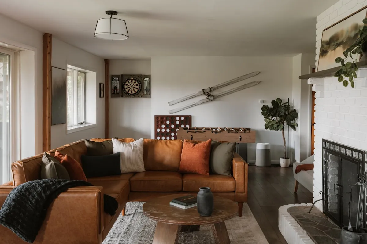 Cozy living room with a tan leather sectional, foosball table, dartboard, and white-brick fireplace with a fiddle-leaf plant