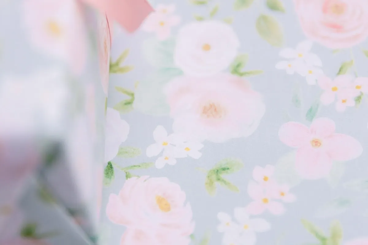 Close-up of a gift wrapped in pastel floral paper printed with watercolor pink roses and white blossoms, tied with a pink ribbon