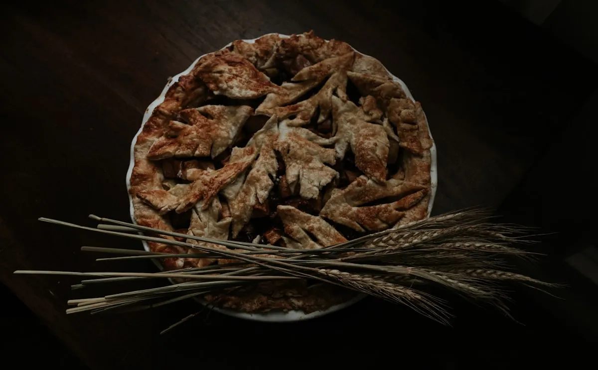 Rustic homemade fruit pie with a decorative leaf-cut crust on a dark wooden table, with stalks of dried wheat across the front