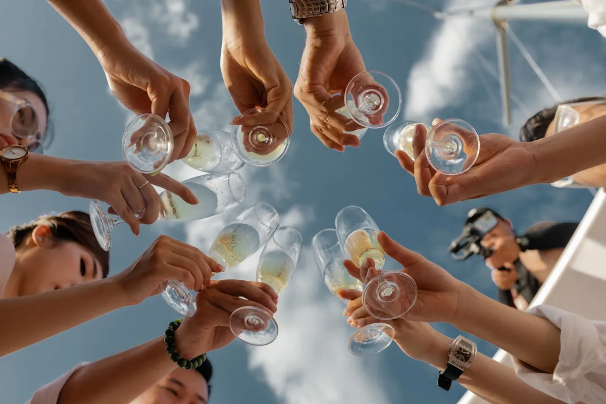 Looking up at a group of friends clinking champagne glasses overhead in a celebratory toast under a bright blue sky