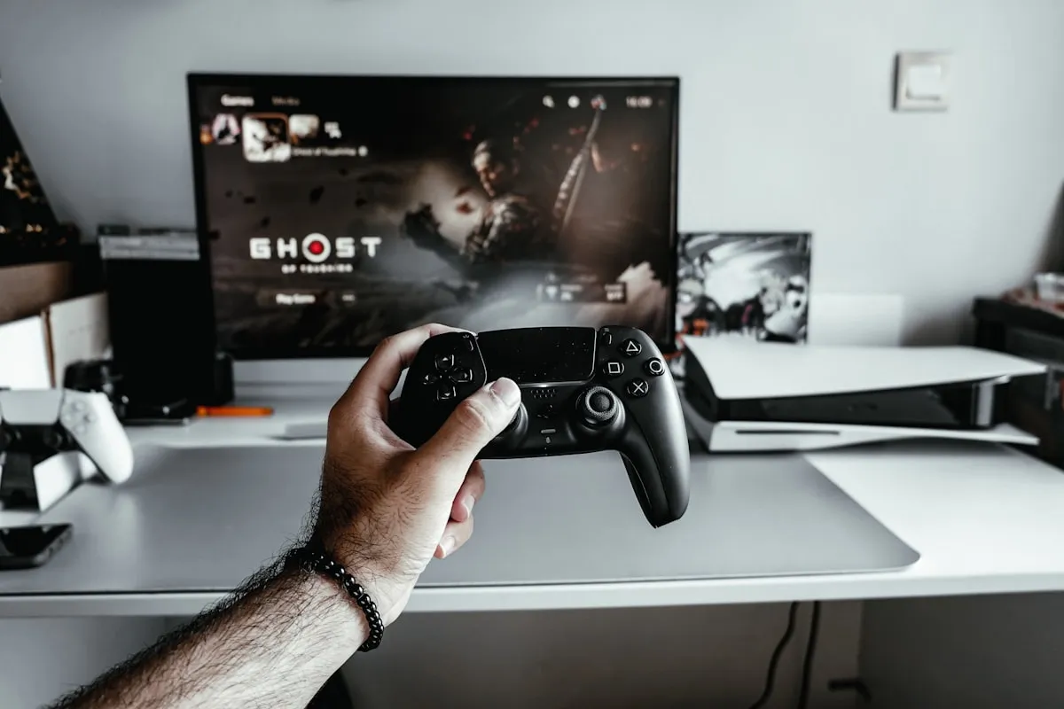 Hand holding a black PlayStation 5 DualSense controller in front of a TV displaying the Ghost of Tsushima game start screen