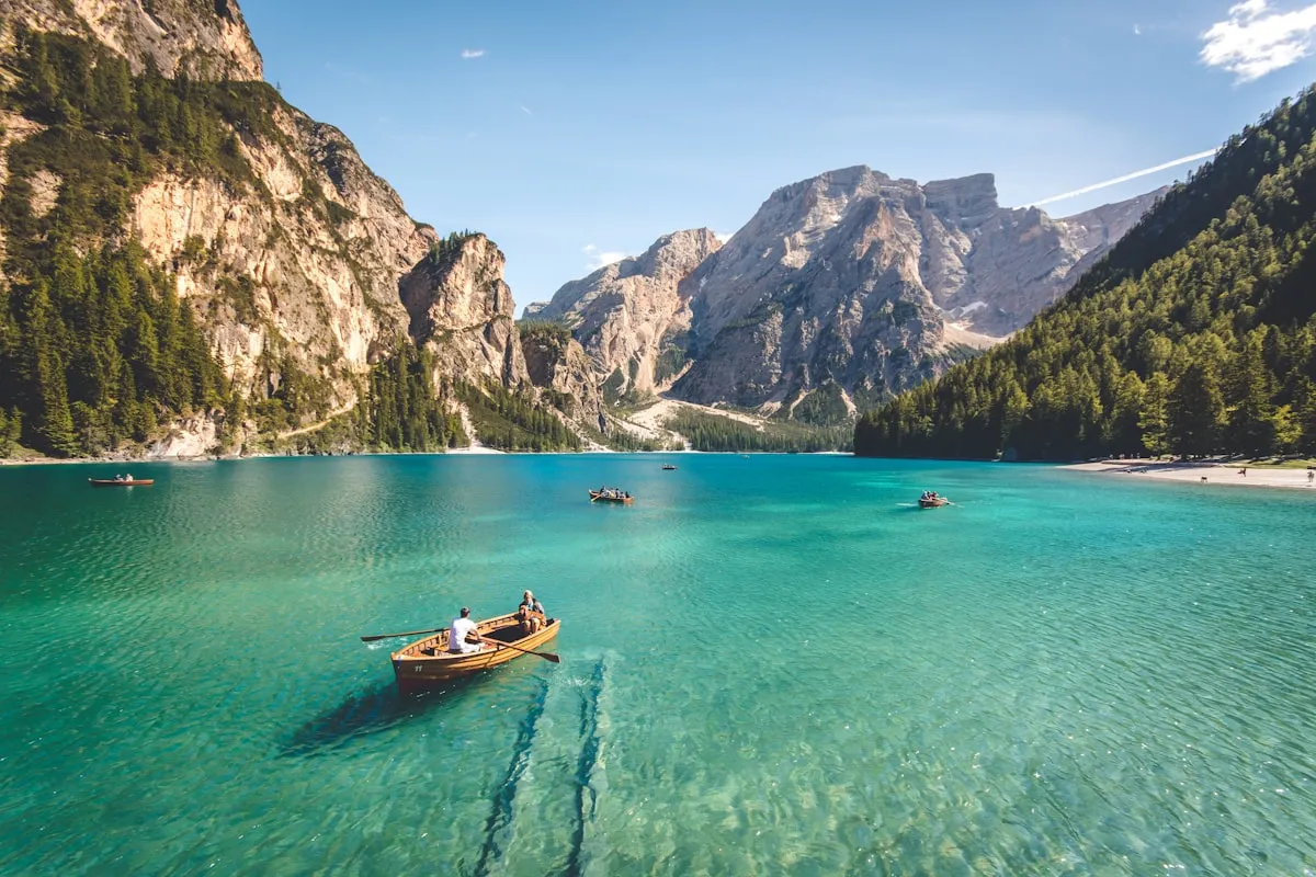 Wooden rowboat with two passengers gliding across a turquoise alpine lake surrounded by tall mountain peaks and pine forest