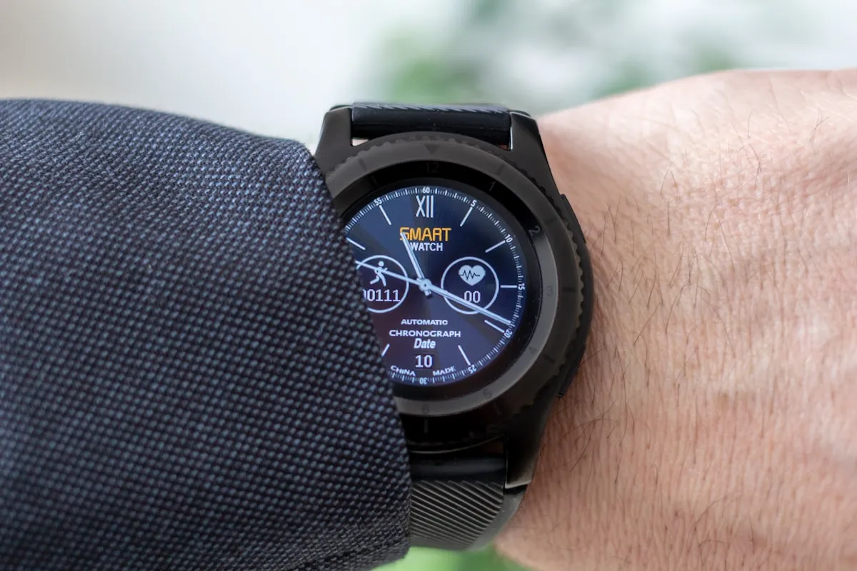 Close-up of a black smartwatch on a wrist showing a chronograph health-tracking face with a heart-rate icon and step count