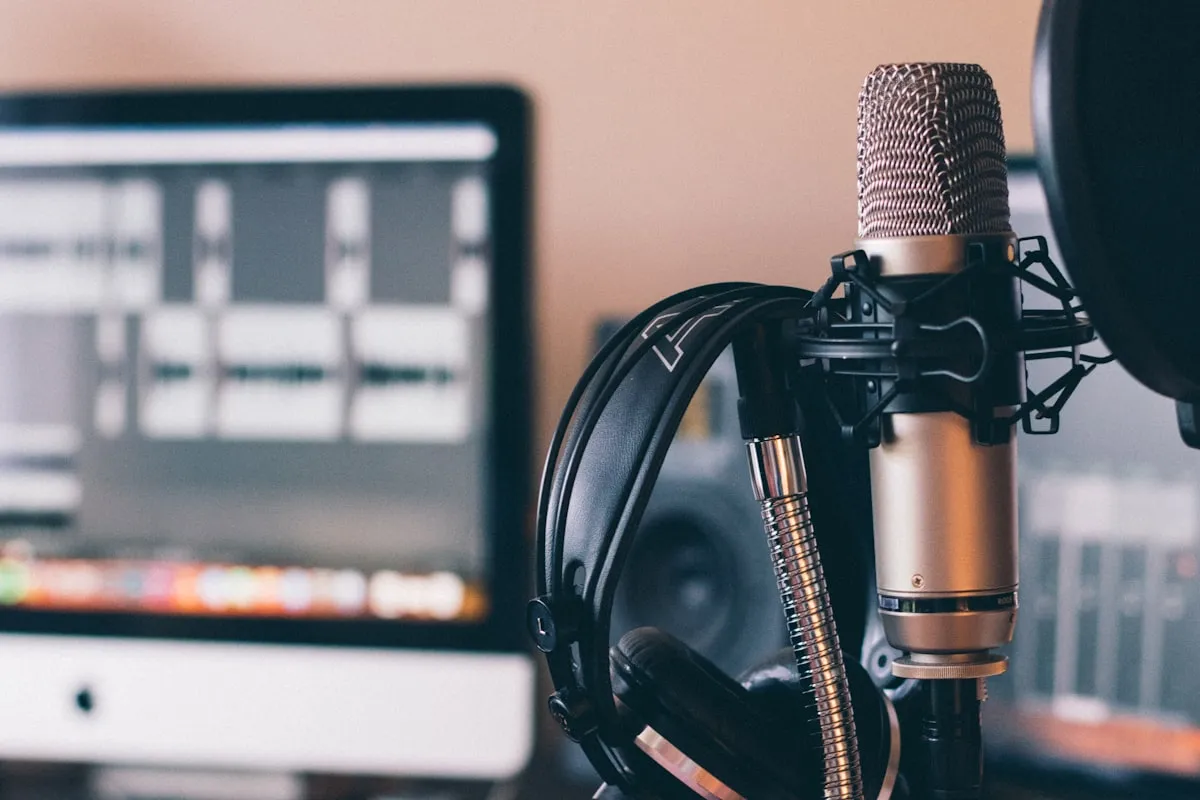 Studio condenser microphone with a pop filter and over-ear headphones in front of a monitor showing a multi-track audio editor
