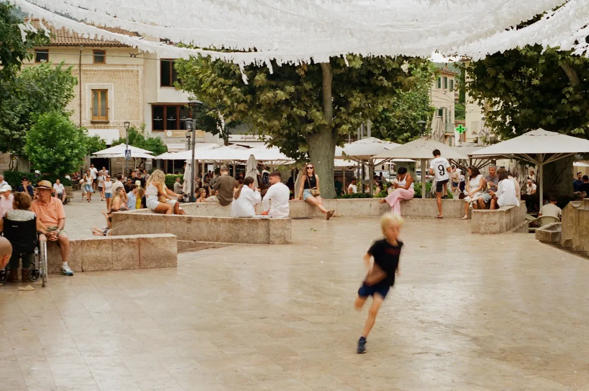 Sunlit town square on a summer afternoon with a child running across the plaza while families and kids gather around fountains and cafes