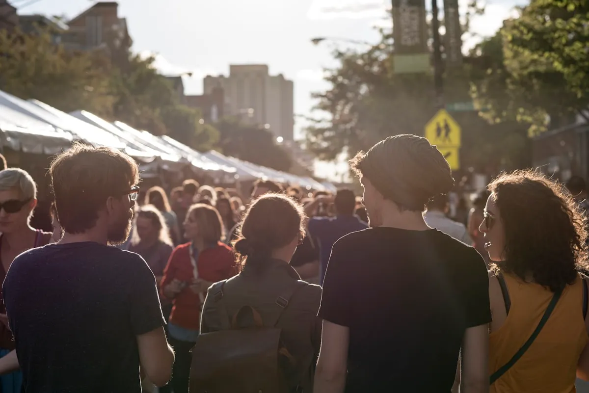 Crowd of shoppers walking through a sunlit outdoor street market past rows of white vendor tents