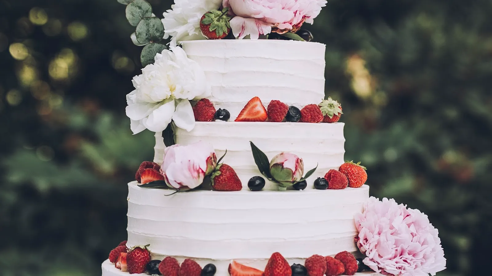 Three-tier white buttercream wedding cake decorated with fresh strawberries, raspberries, blueberries, and pink and white peony blooms
