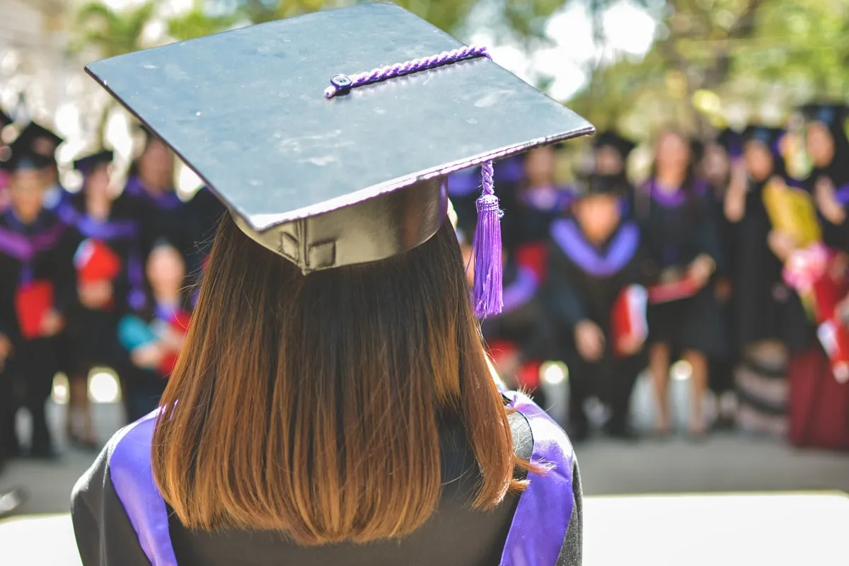 Back view of a college graduate in a black mortarboard cap with a purple tassel, facing a crowd of fellow graduates at a ceremony