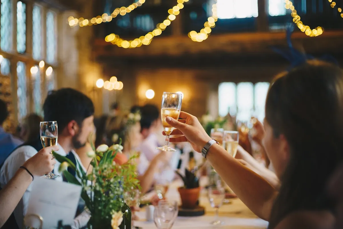 Wedding guests at a long reception table raising champagne flutes for a toast under warm string lights