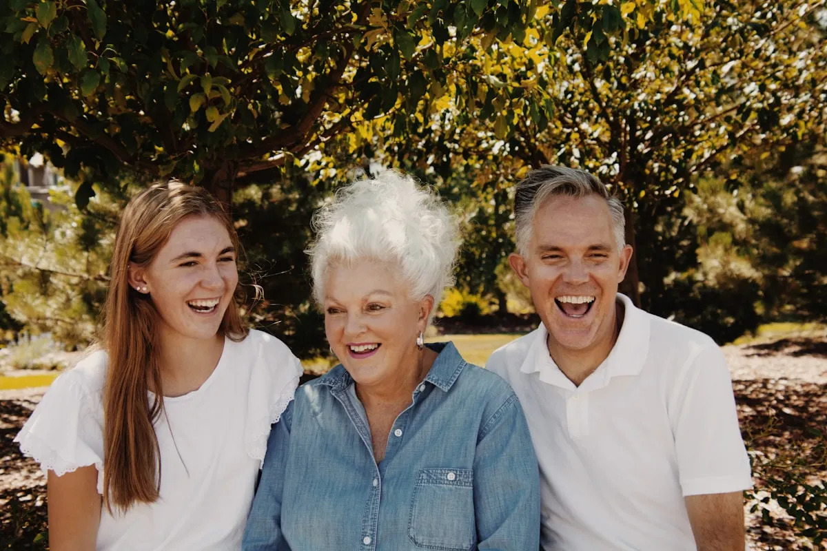 Adult son and adult daughter laughing on either side of their senior mother under a sunlit tree, all wearing white and denim