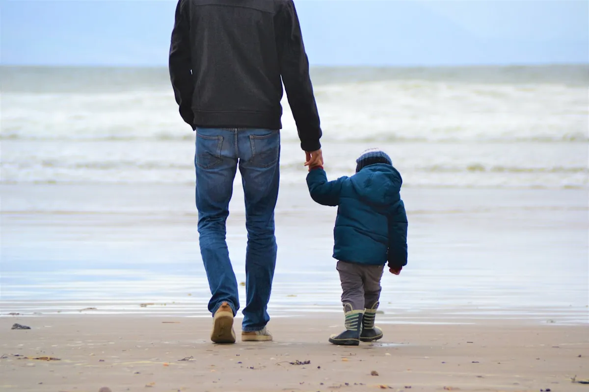 A father holding the hand of his small child as the two walk together along a sandy beach toward the ocean on an overcast day