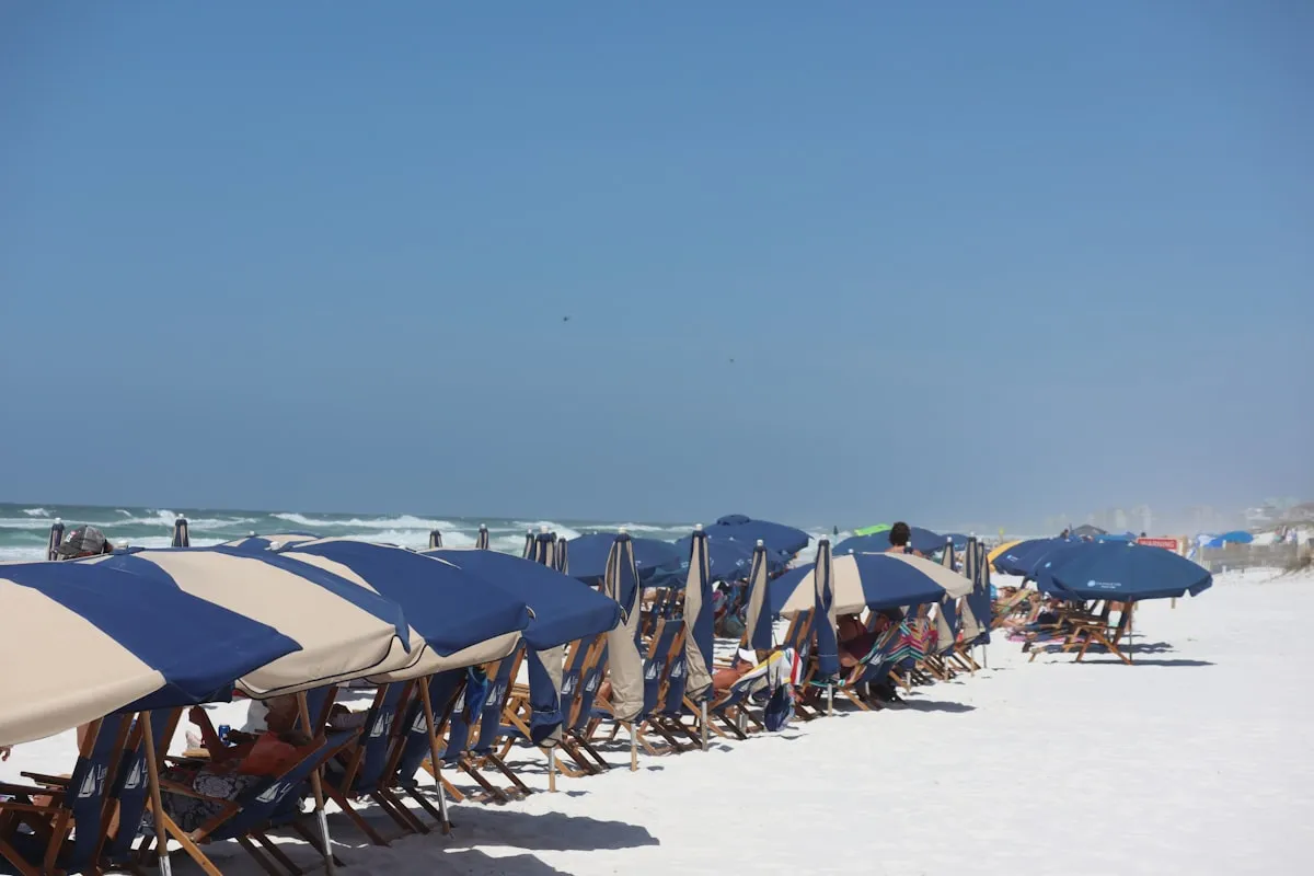 A row of blue and white striped beach umbrellas and rental chairs lined up on white sand, with families relaxing under them and the ocean visible in the background