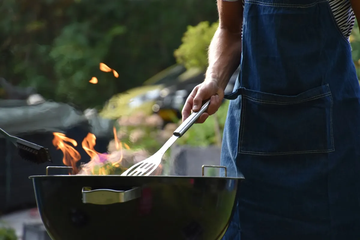A man in a denim apron tending a kettle grill with bright flames rising from the coals in a backyard — the signature scene of Memorial Day weekend