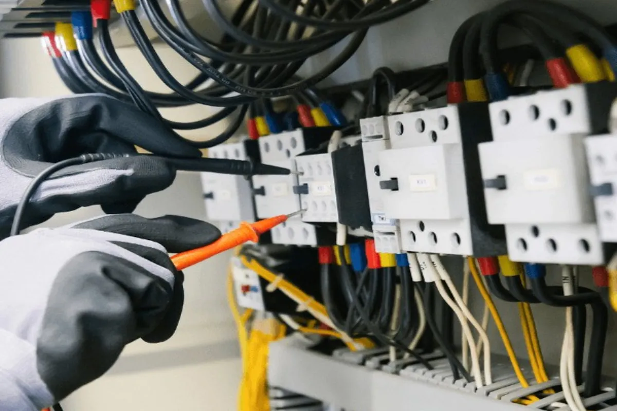 Gloved hands holding black and orange test probes against a row of white contactors and red, yellow and blue crimped wire ferrules inside an industrial electrical control panel