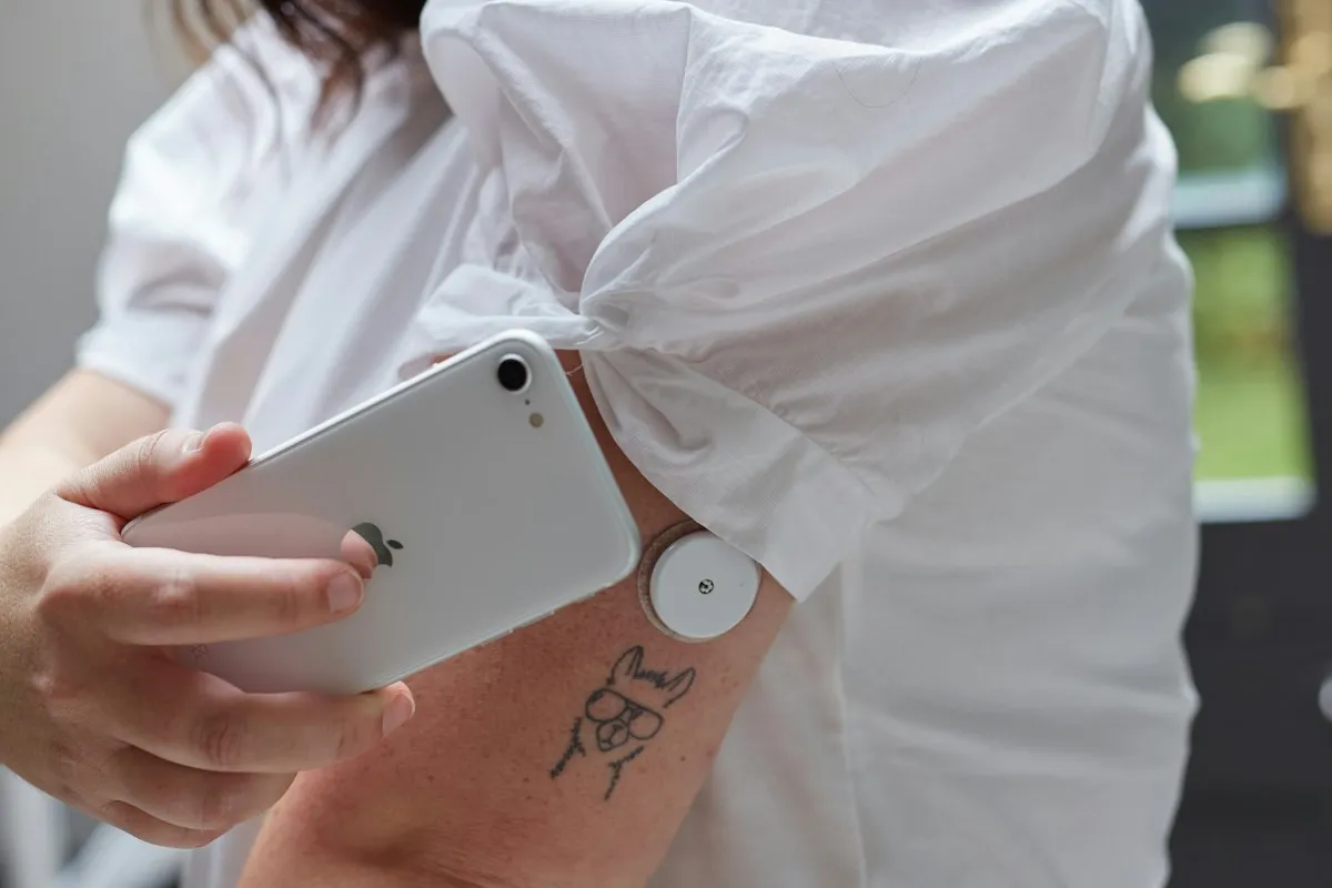 A person in a white shirt with a rolled-up sleeve holds the back of a white smartphone against a round glucose sensor worn on their upper arm, just above a small llama tattoo.