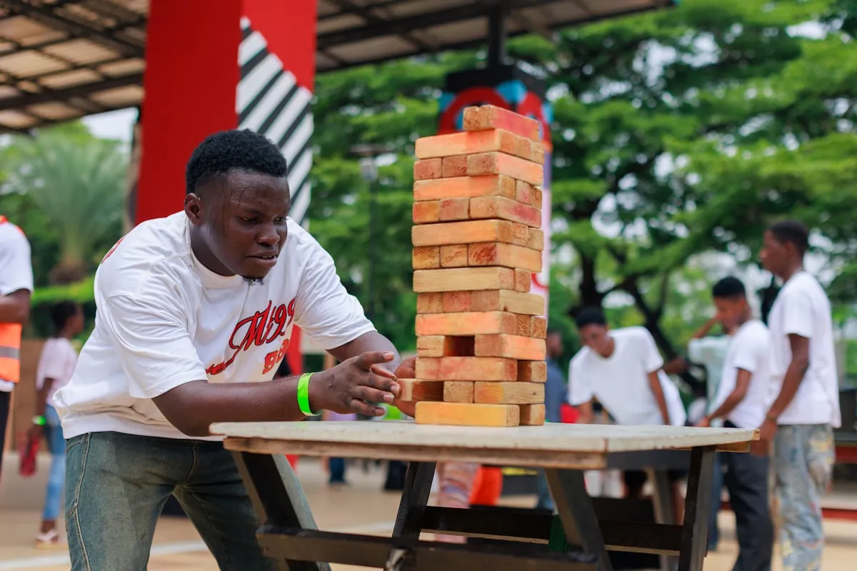 A young man in a white T-shirt carefully sliding a wooden block out of a tall, precarious giant Jenga tower on an outdoor table, with other people in the background