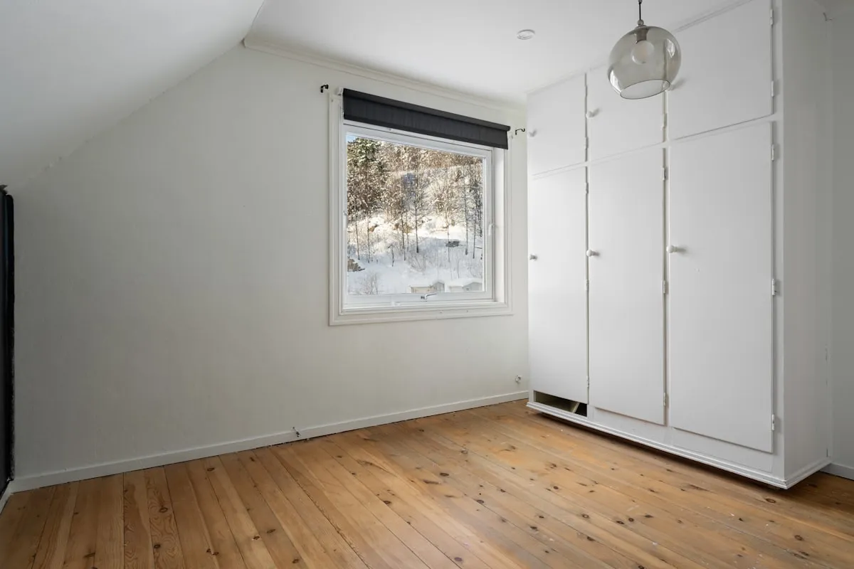 Empty apartment room with white walls, a sloped ceiling, a built-in white wardrobe, wide wooden plank flooring, and a window looking out onto a snowy wooded hillside