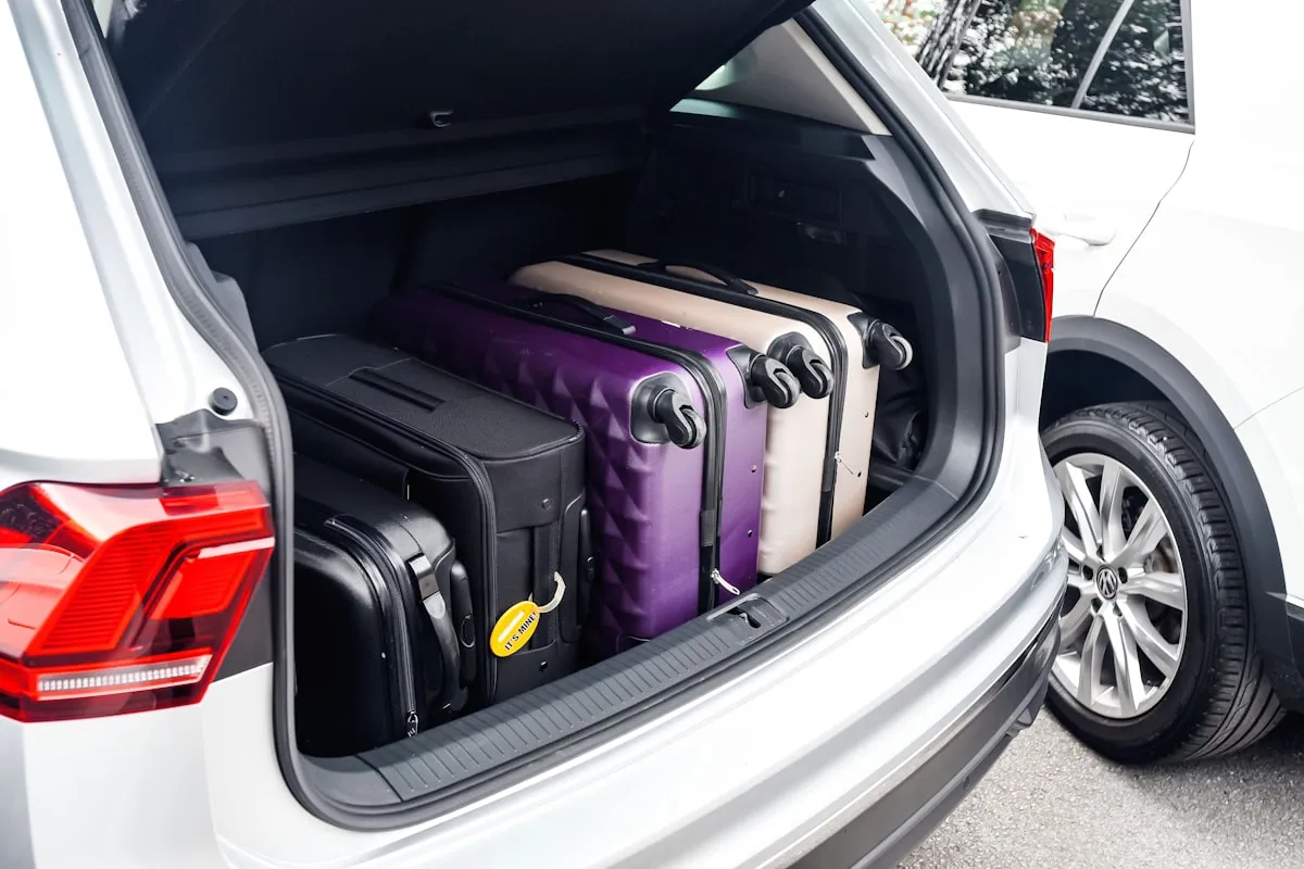 Four hard-shell suitcases in black, purple and cream packed tightly into the open trunk of a white SUV parked on a driveway