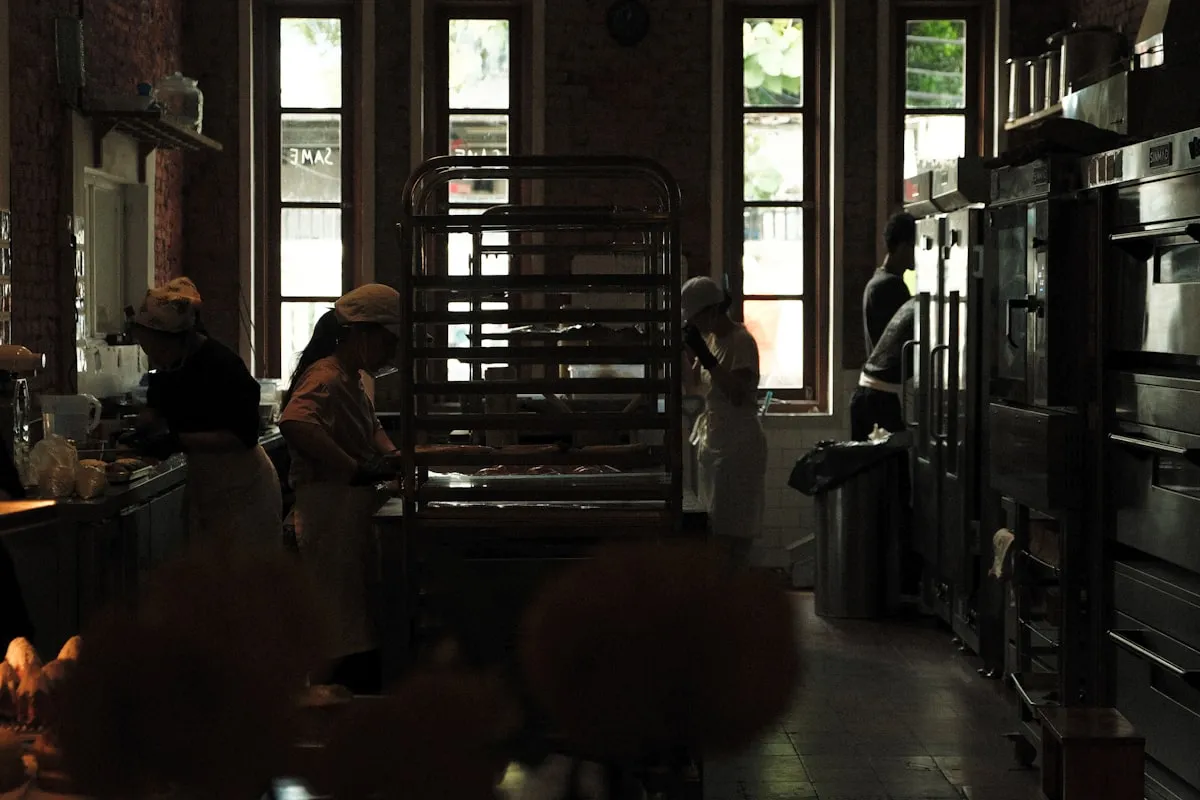 Four bakers in aprons and caps working at separate stations in a dim brick-walled bakery, with a tall sheet-pan rack and ovens between them