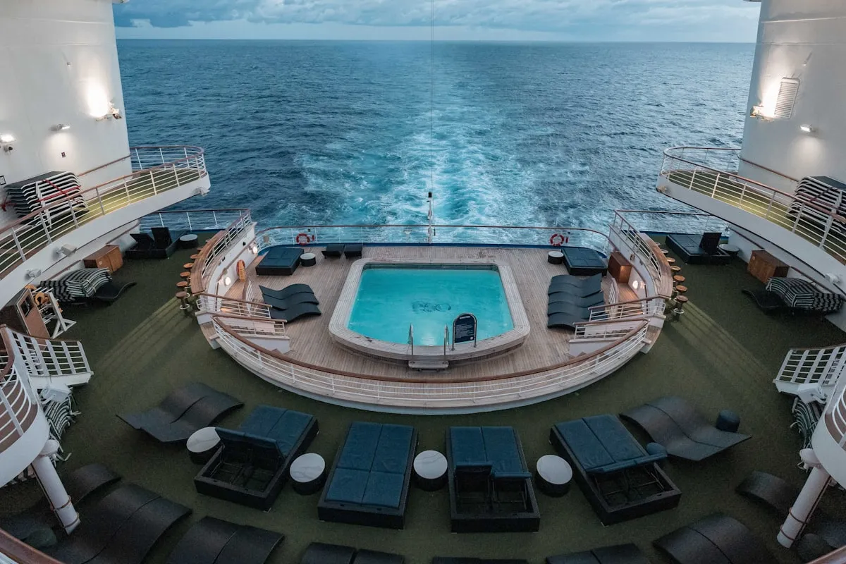 Aerial view of a cruise ship's stern pool deck at dusk — a small turquoise pool ringed by empty black loungers on a wooden deck, overlooking the ship's white wake across a gray ocean