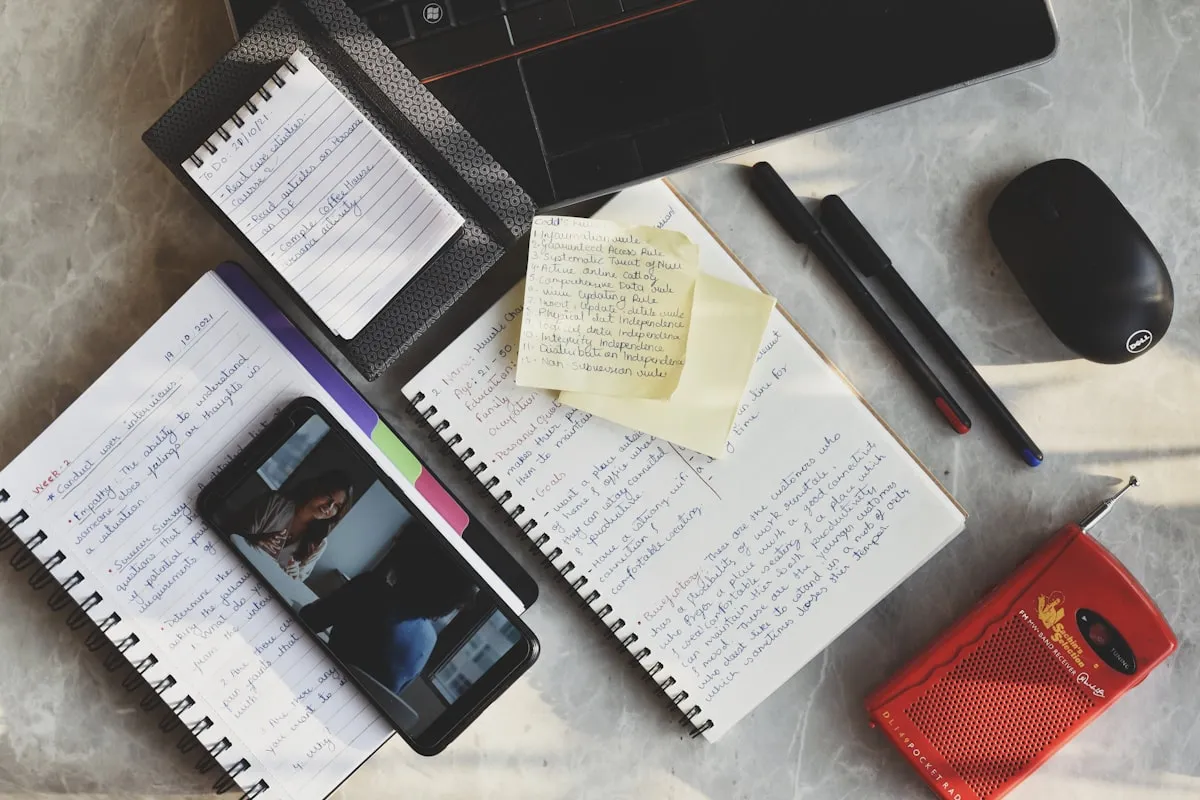 Overhead view of a cluttered desk with a laptop edge, spiral notebooks full of handwritten notes, yellow sticky notes, pens, a computer mouse, a smartphone showing a photo, and a small red radio